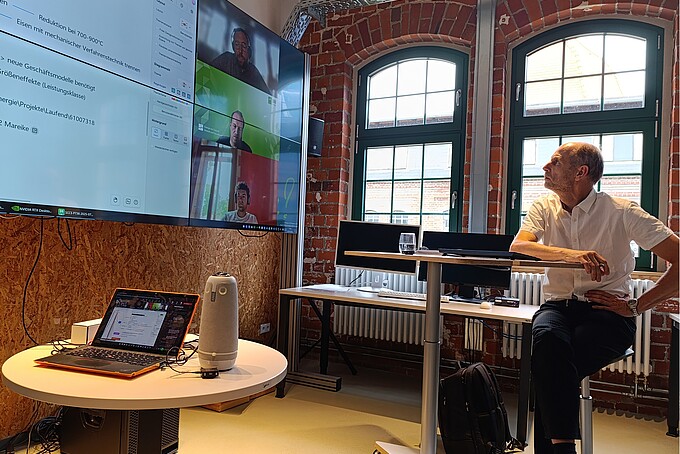 Professor Zschunke sitzt auf einem Hochstuhl und schaut auf eine große Videoleinwand in einem Arbeitsraum mit Backsteinwänden und Halbrundfenstern.