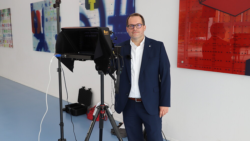 Der Rektor steht im Foyer von Haus vier neben einem Teleprompter und lächelt in die Kamera.