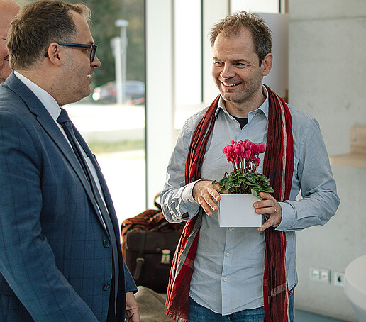 Der Rektor unterhält sich mit einem Professor, der eine Topfpflanze in den Händen hält.