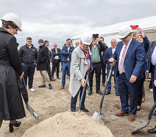 Der Rektor hält eine Schaufel in der Hand und nimmt eine Schippe Sand auf. Neben ihm tun es ihm die anderen Projektbegleitenden gleich. Im Hintergrund stehen Gäste der Veranstaltung.