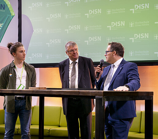 Moderator Emil, Prof. Hänsel und der Rektor stehen auf dem Podium.