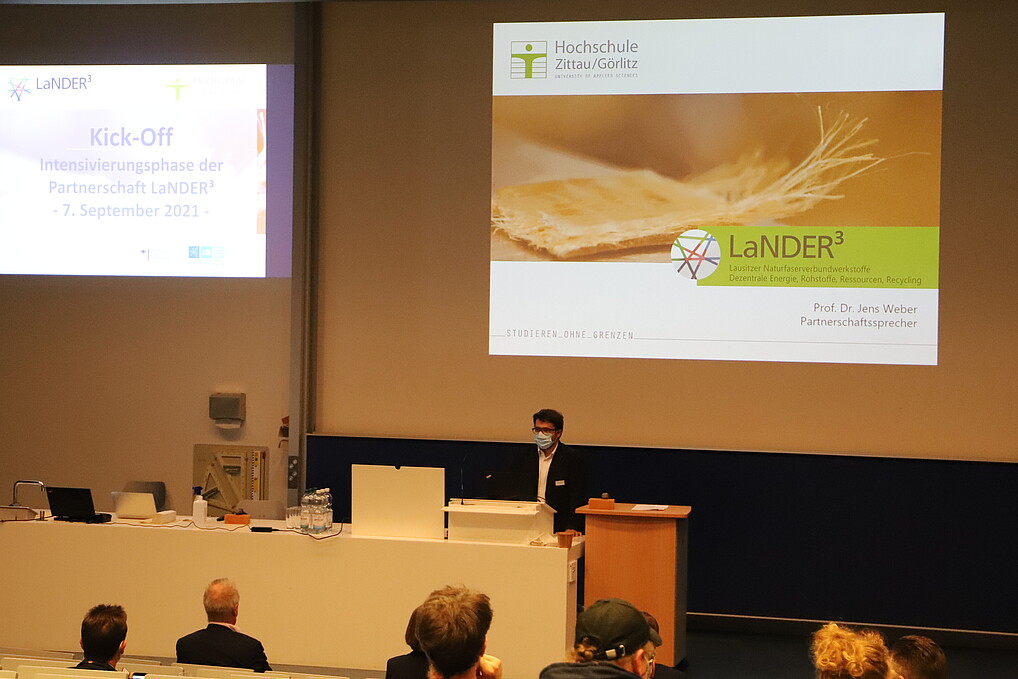 Im Vorlesungssaal hält ein Professor eine Rede. Im Hintergrund wird das Programm projiziert.