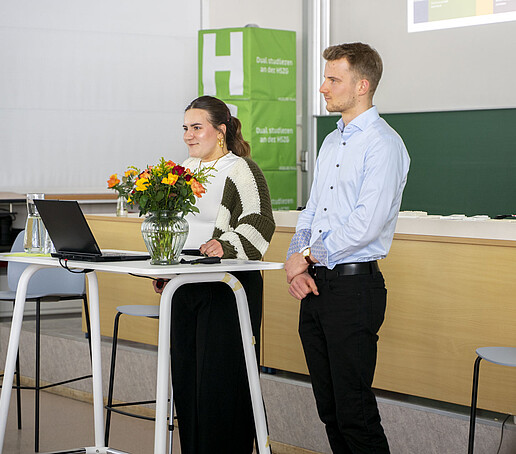 Ein Mann und eine Frau stehen hinter einem Tisch im sind dem Publikum zugewandt.