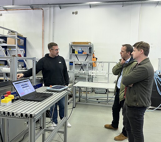 Ein Mann erklärt 2 anderen Personen die Cobot Versuchslinie