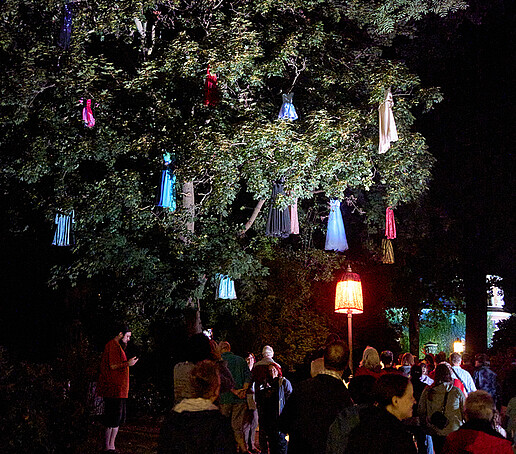 Bunte Tücher hängen in einem Baum, Besucher laufen darunter durch.
