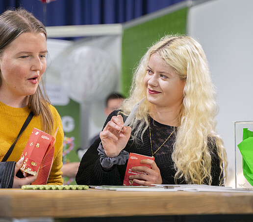 Zwei junge Studieninteressierte mit Popcorn-Tüten stehen an einem Holztisch.
