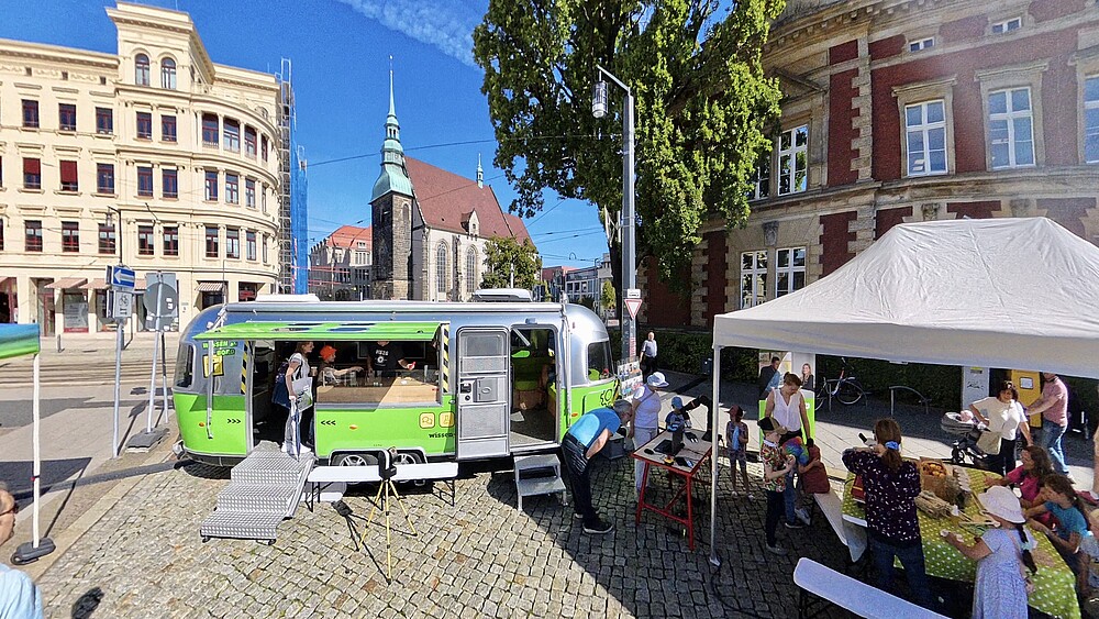 Der Mobile Lernort-Anhänger steht mit offenen Türen und Seitenfenster auf dem Görlitzer Postplatz. Daneben steht ein weißer Pavillon. Darunter beschäftigen sich Kinder und andere Gäste mit den ausgelegten Materialien.