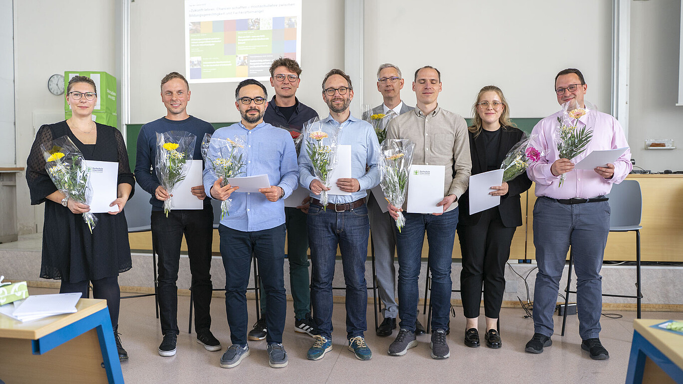Eine Gruppe von Hochschulmitarbeitenden steht nebeneinander. Sie halten eine Blume und eine Urkunde in den Händen.