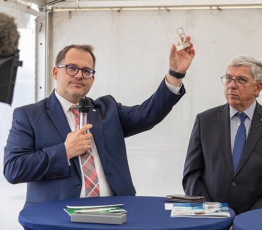 Der Rektor der HSZG hält ein HSZG-Signet aus Metall in der Hand an einem Rednerpult in einem Zelt. Neben ihm steht ein Mann.
