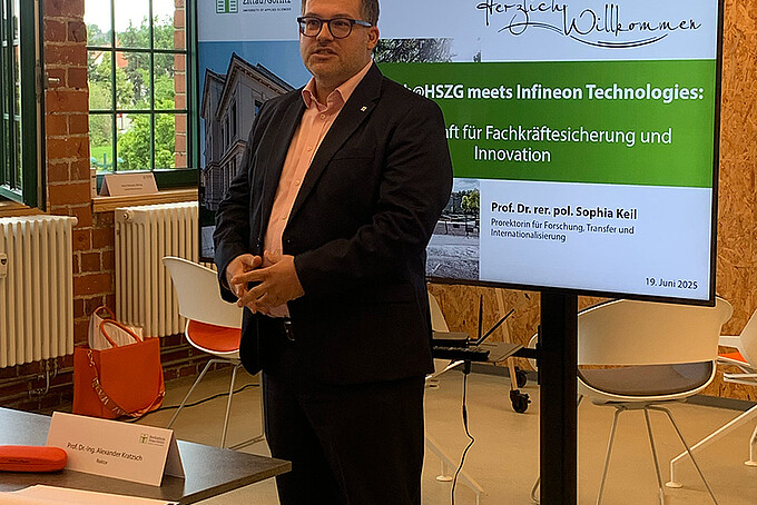 Der Rektor hält Grußworte vor einem Bildschirm.