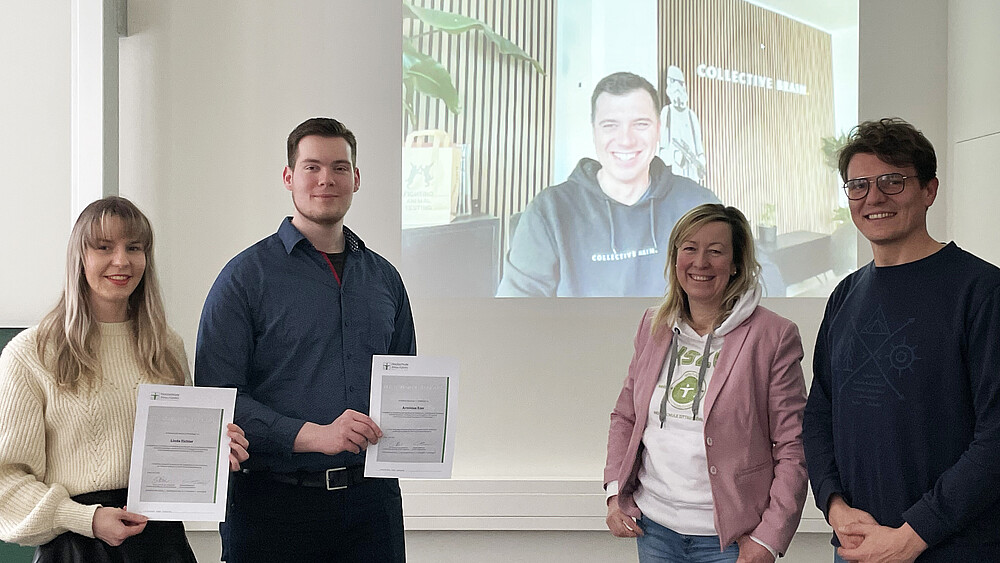 Das Zuklos-Team und Studierende stehen vor einer Wand, auf die ein Agenturmitarbeiter per Webcamübertragung projiziert ist.