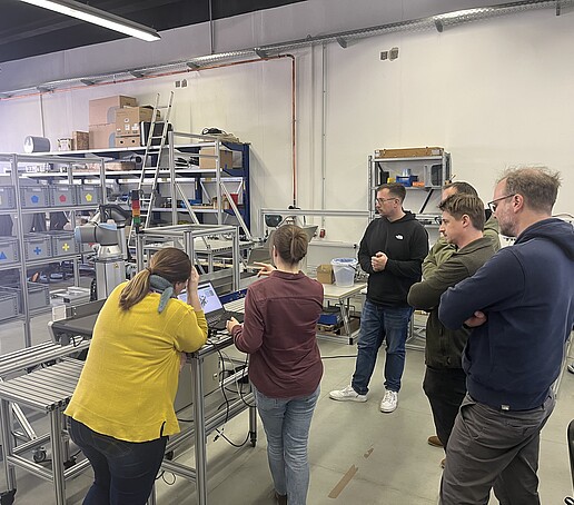 Mehrere Personen schauen sich die Ein Mann erklärt 2 anderen Personen die Cobot Versuchslinie an