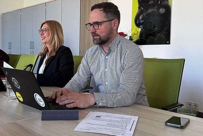 Eine Professorin und ein Professor sitzen in der Kommissionssitzung, er tippt in seinen Laptop, sie lächelt in die Runde.