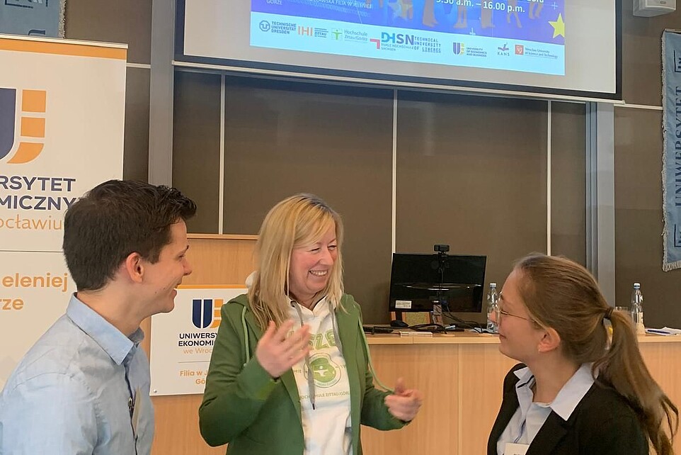 Die Prorektorin im Gespräch mit zwei Nachwuchsforschenden der HSZG bei der Konferenz. Im Hintergrund leuchtet eine Präsentation auf einer Leinwand.