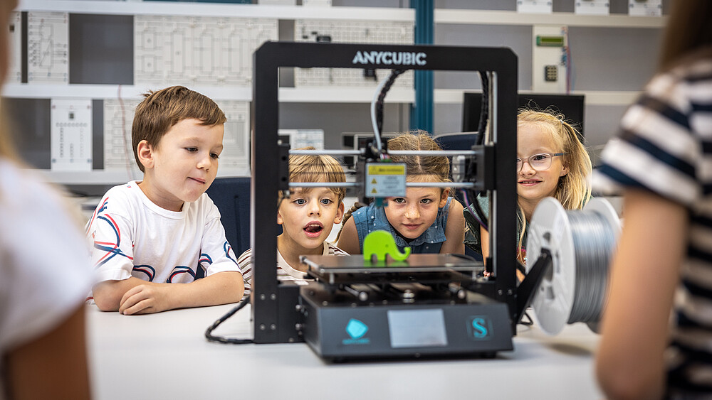 Vier Kinder sitzen im Elektrotechnik-Laborsaal und schauen sich einen kleinen, grünen Elefanten auf einer Plattform eines Gerätes an.
