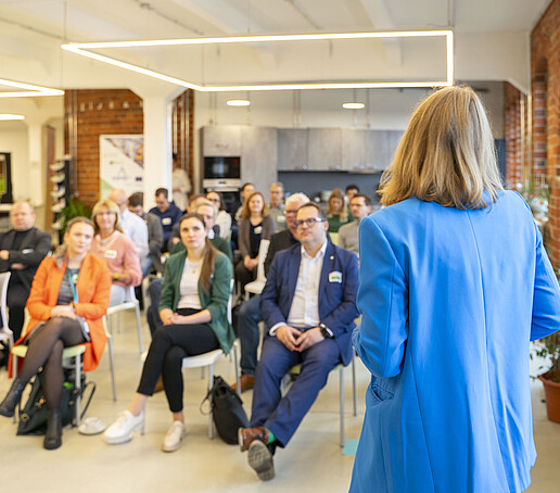 Die Moderatorin ist dem Publikum zugewandt, dass auf Stuhlreihen sitzt.