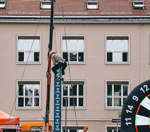 Beim Bierkastenstapeln steht eine Person weit oben und ist über ein Hubfahrzeug gesichert. Ein Mitarbeiter mit Helm steht daneben und sichert den Kletterer ab.