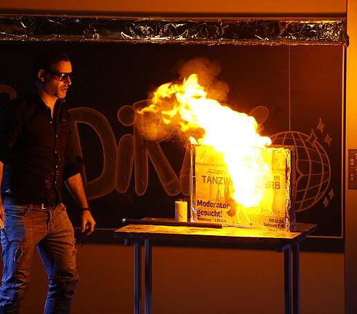 Eine Person führt im Dunkeln ein flammendes Experiment auf der Bühne durch.