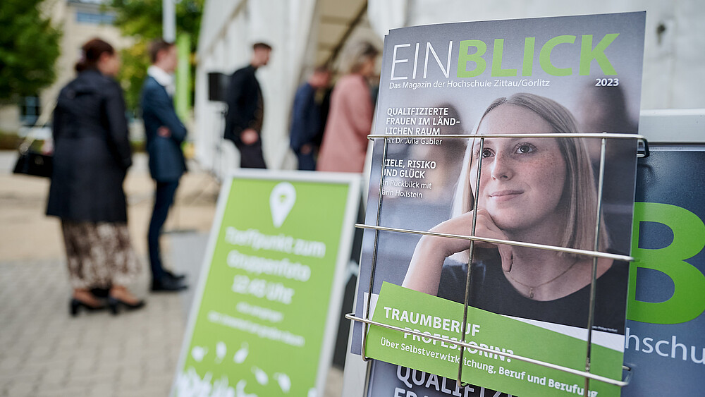 Ein Aufsteller mit Körbchen und Einblick-Magazinen darin. Im Hintergrund gehen Menschen in ein großes Zelt.