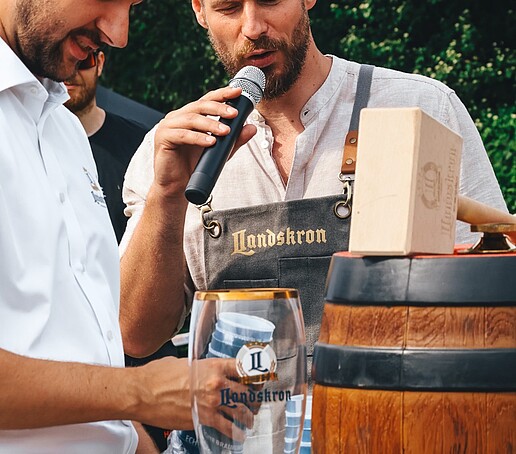 Der Hochschulsportzentrumsleiter spricht in ein Mikro, während ein junger Mann ein Bierfass präpariert-.