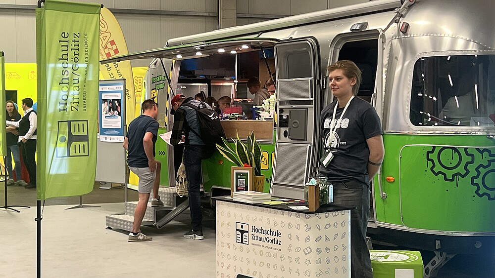 Der Airstream-Anhänger steht in der Messehalle und Besuchende betreten ihn und schauen sich Experimente an. Im Vordergrund steht eine Mitarbeiterin an einer HSZG-Theke.