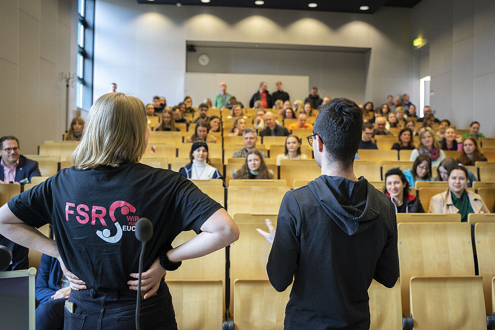 Zwei Studierende, Moderator Lennard und Sahra von der Fachschaft Sozialwissenschaften, stehen vor sitzenden Studieninteressierten im Hörsaal und informieren über das Studienangebot und das Campusleben an der Hochschule Zittau/Görlitz. Das Bild zeigt die offene und kommunikative Art der Hochschule, Studieninteressierte willkommen zu heißen und bei ihrer Entscheidungsfindung zu unterstützen. Es vermittelt eine lebendige Atmosphäre und zeigt, wie engagiert und begeistert Studierende und Mitarbeiter der Hochschule sind, Interessenten für ein Studium an der Hochschule Zittau/Görlitz zu begeistern.