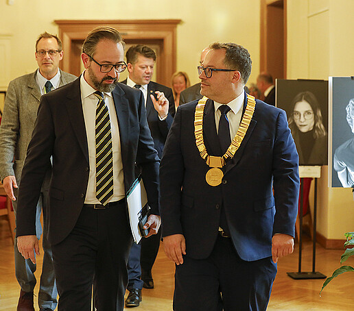 Wissenschaftsminister und Rektor gehen in das Foyer neben dem Ratssaal in dem eine Fotoausstellung aufgebaut ist.