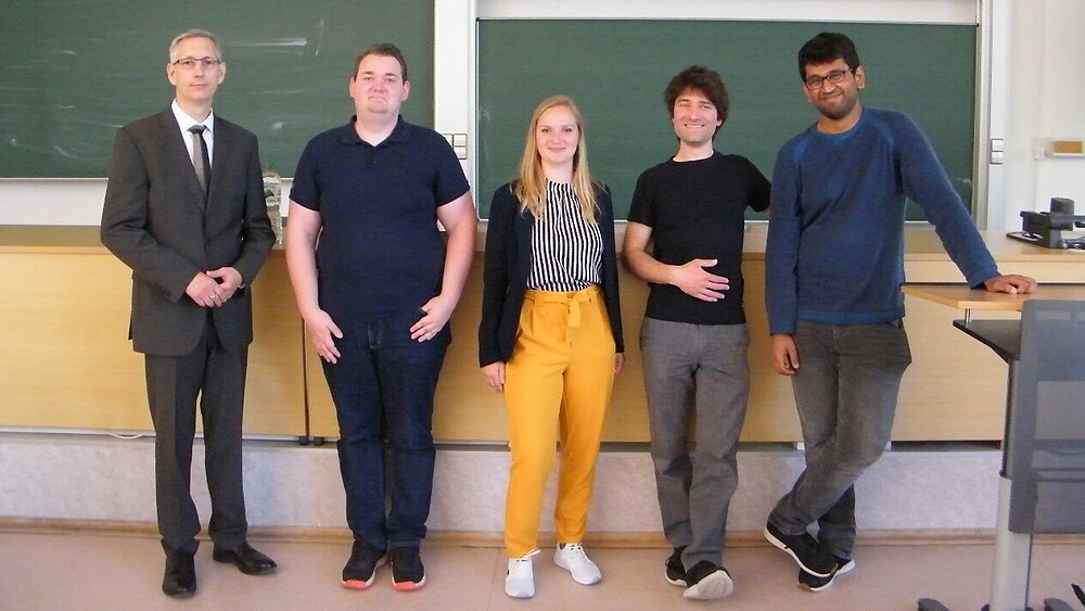 Im Hörsaal stehen fünf Personen vor den Tafelboards und lächeln in die Kamera: Prof. Bellair, Herr Eik-Fabian Gleiß, Frau Johanna Schubert, Herr Sven Wiedemann, Herr Rajesh Sriram. 