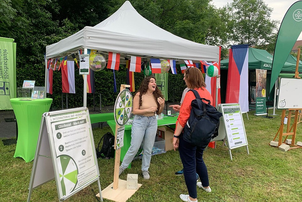 Festteilnehmende unterhalten sich vor dem International Day Pavillon. Davor steht ein Glücksrad. An dem Pavillon hängen internationale Flaggen.