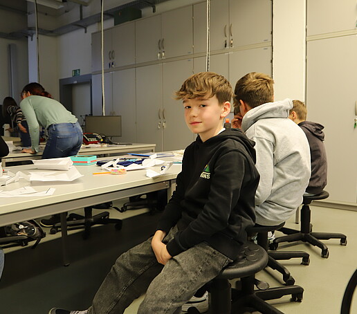 Ein Schüler sitzt im Werkstofflabor am Tisch und lächelt in die Kamera.