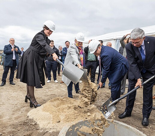 Der Rektor und die anderen Projektbeteiligten werfen jeweils eine Schippe Sand in ein Erdloch. Im Hintergrund stehen Gäste der Veranstaltung.