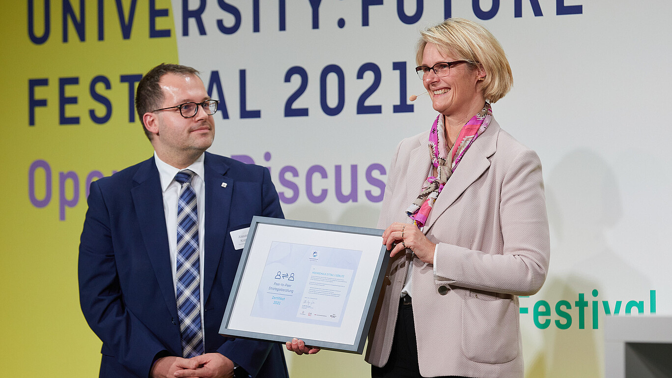 Bundesbildungsministerin Anja Karliczek übergibt dem Rektor der HSZG eine Urkunde und lächelt in die Kamera.