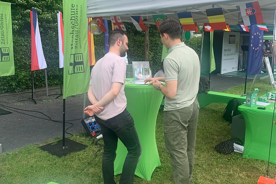Zwei Männer stehen am grünen Stehtisch und schreiben eine Postkarte.