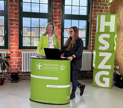 Eine Mitwirkende und die Moderatorin stehen im Studio hinter einem HSZG-Theke. Links im Vordergrund steht eine schwarze Kamera. Rechts von der Theke sind vier HSZG-Würfel aufgetürmt.