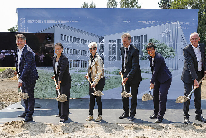Initiatoren des DLR-Institutes halten jeweils einen mit Erde gefüllten Spaten vor sich und schauen in die Kamera. Im Hintergrund ist eine Pressewand aufgebaut, auf dem der Entwurf des künftigen Institutes abgebildet ist.