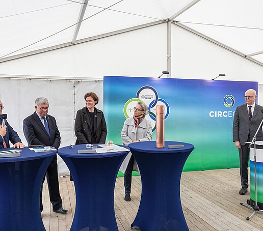 Die Beteiligten am CircEcon-Projekt stehen in einem weißen Zelt an blauen Stehtischen und unterhalten sich vor einer blau-grünen Pressewand, auf der das CircEcon-Logo aufgedruckt ist.