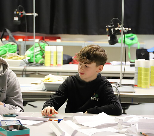 Ein Schüler sitzt am Tisch und schaut auf die Bastelsachen seines Tischnachbarn. 