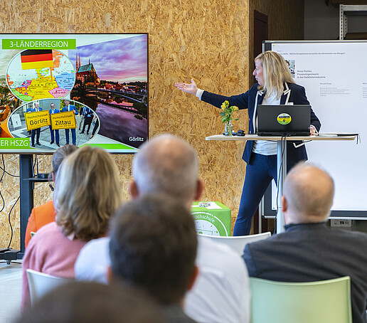 Die Prorektorin hält am Stehtisch einen Vortrag und deutet auf einen Bildschirm.