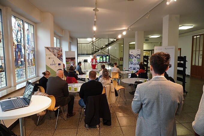 Die Prorektorin spricht im Hintergrund nach vorn gewandt zum Publikum, das an Rundtischen sitzt.