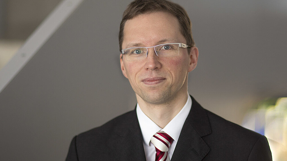 Stefan Kornhuber im Anzug mit Krawatte und Brille lächelt in die Kamera.