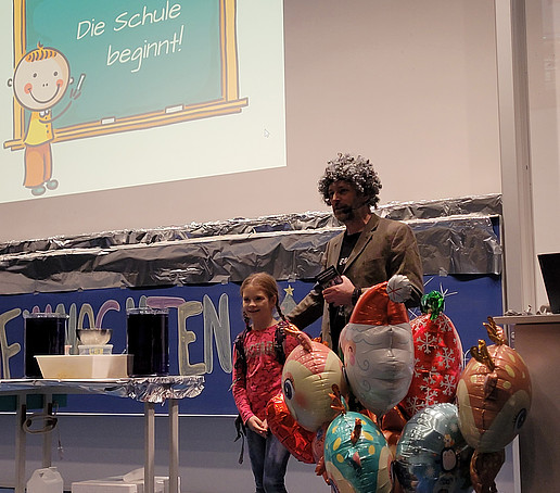 Ein Mädchen und ein Mann mit Perücke stehen auf der Bühne, im Vordergrund steht ein Behälter mit Luftballons.