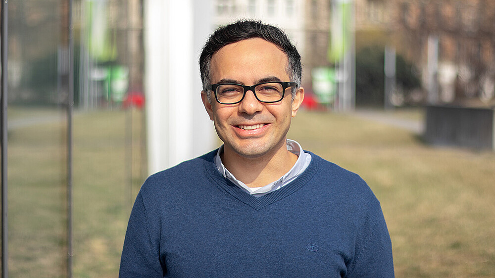 Prof. Massoudi im blauen Pulli und mit Brille steht im Freien neben einer Glasfassade und lächelt in die Kamera.