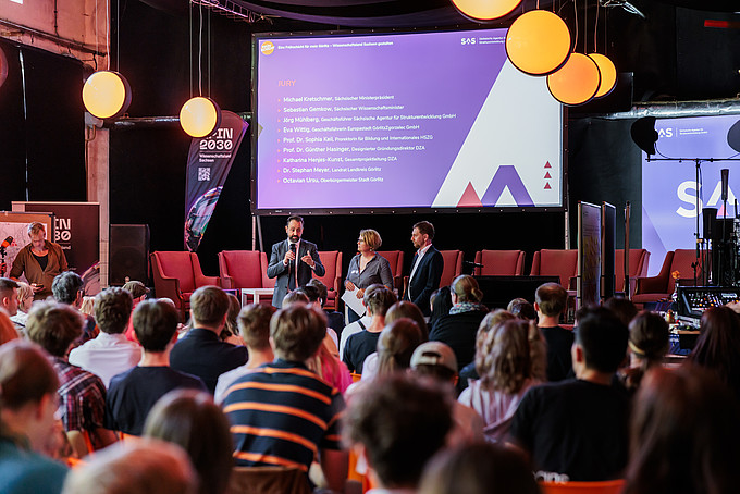 Sprecher*innen auf der Bühne während der Präsentation vor einer großen Leinwand.