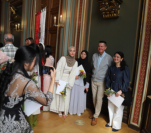 Ein Dozent posiert mit einer Gruppe von Absolventinnen neben der Zittauer Fahne im Bürgersaal.