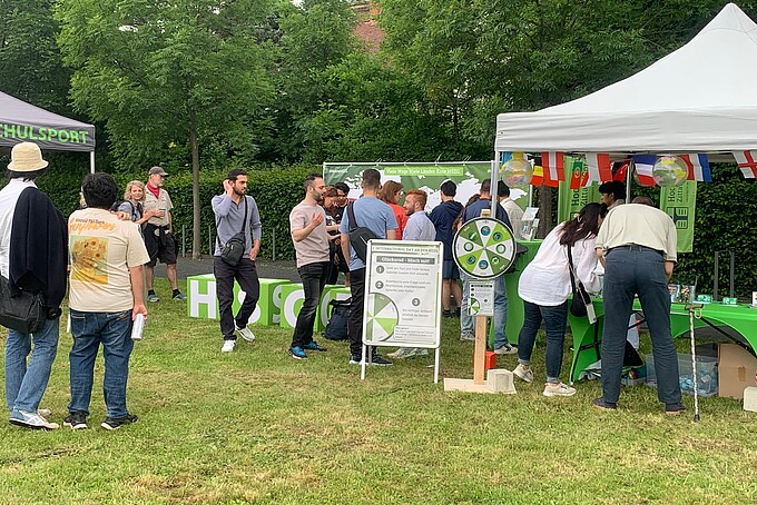 Festteilnehmende unterhalten sich vor dem International Day Pavillon. Davor steht ein Glücksrad. An dem Pavillon hängen internationale Flaggen.