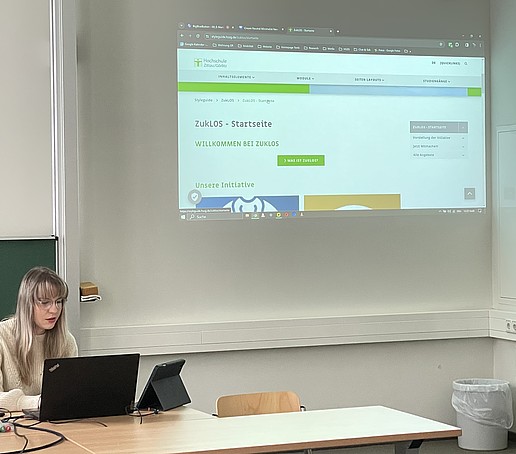 Eine Studentin sitzt am Tisch hinter einem Laptop und hält eine Präsentation.