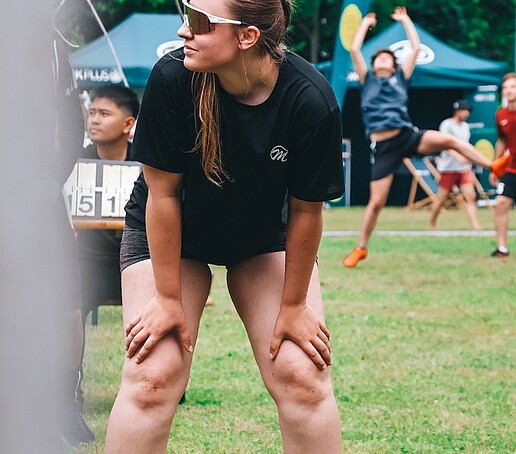 Eine Studentin steht auf dem Wiesensportplatz und stützt ihre Hände auf ihre Knie.
