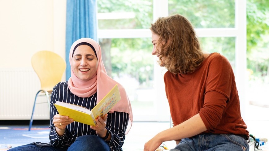 Zwei junge Frauen sitzen in einem Seminarsaal auf dem Boden, die linke Frau hält ein aufgeschlagenes Buch in der Hand. Beide lachen.
