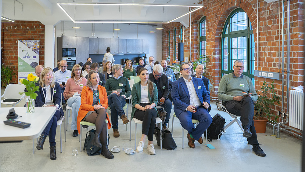 Workshop-Teilnehmende sitzen auf Stühlen im einem Raum mit roten Steinwänden und richten ihren Blick in Richtung Vortragenden.