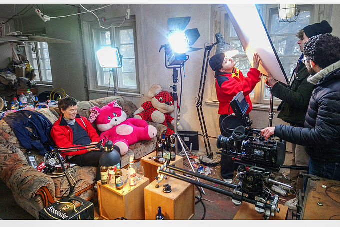 Dreharbeiten zum SFA Abschlussfilm „Katharina“. Kursteilnehmende überprüfen die Setbeleuchtung, während eine Kursteilnehmerin in der Kulisse auf einem alten Sofa sitzt, umgeben von pinken Plüschtieren und leeren Bierflaschen. 